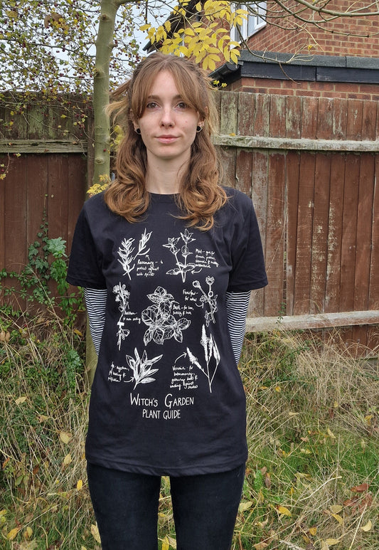A ginger woman wears a black t-shirt with a white botanical plants illustration featuring the words 'Witch's Plant Guide' at the bottom. She wears a black and white striped top with the t-shirt as well as black jeans.