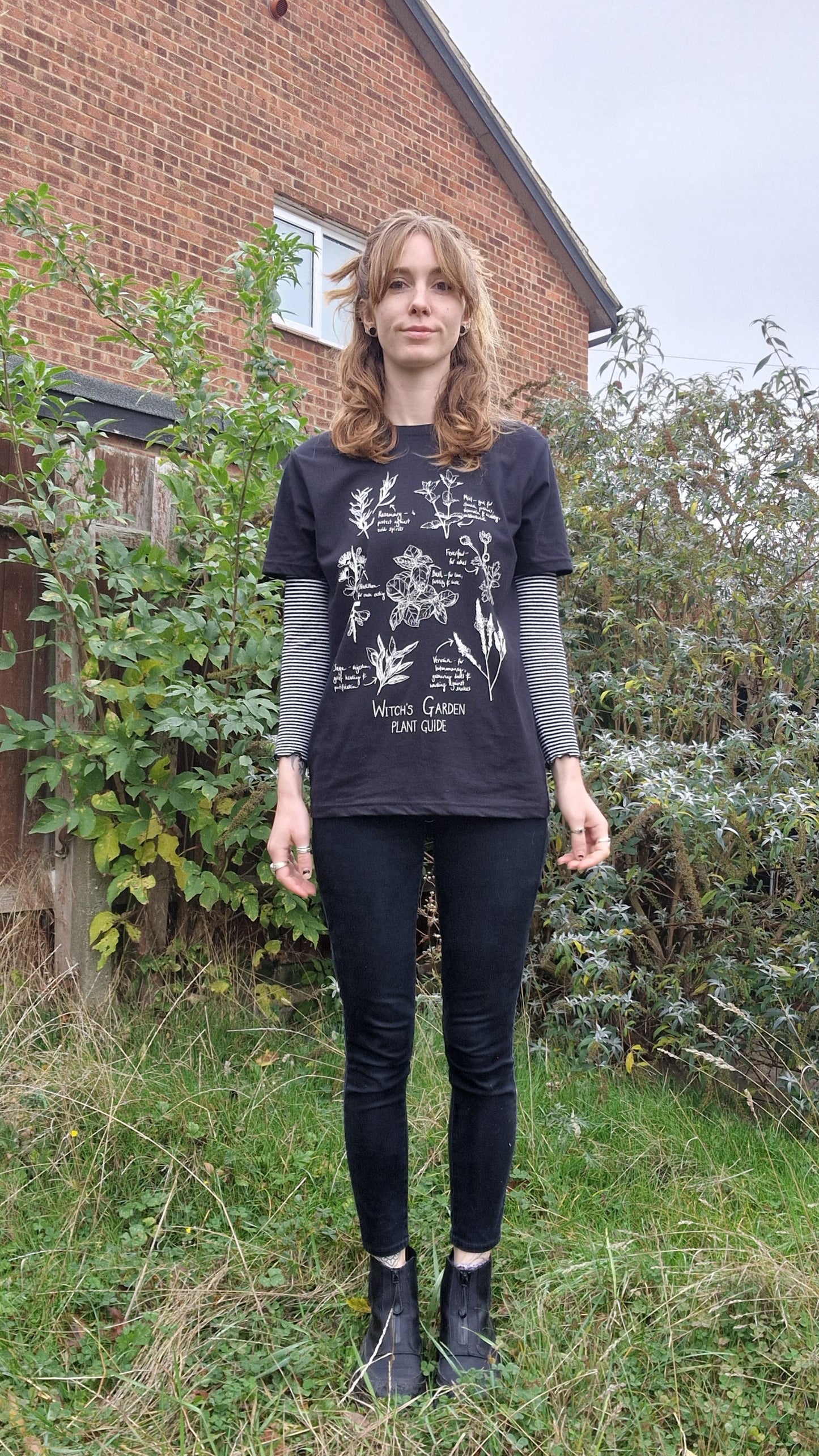 A ginger woman wearing a black tshirt with a white design of plants over the words 'Witch's Garden Plant Guide'. She stands in an overgrown garden, and wears the t-shirt with a striped white and black long sleeved top, black jeans and black boots.
