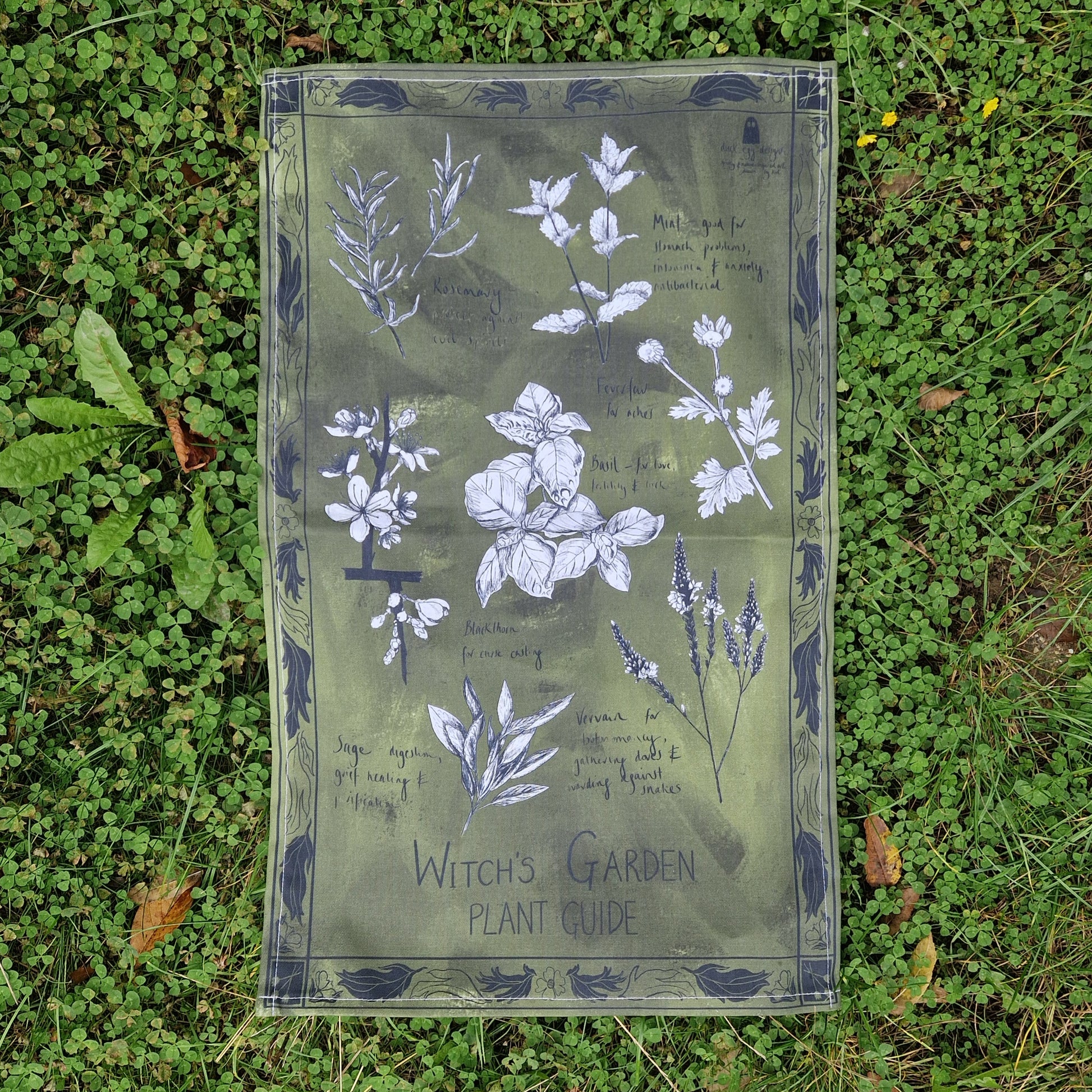 A mottled green tea towel rests on a bed of vibrant green clover. The tea towel features a black leafy border and the words 'Witch's Garden Plant Guide' with seven different black and white botanical plant illustrations and their uses. 