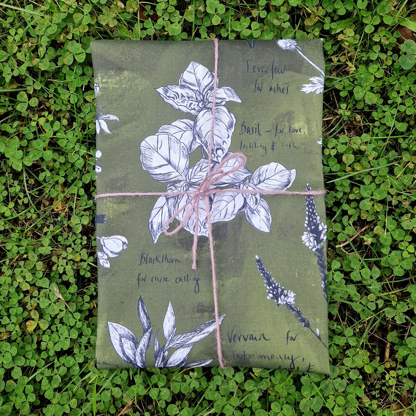 A folded green tea towel with white and black botanical plant illustrations, wrapped in lilac twine. The tea towel rests on a patch of green clover.