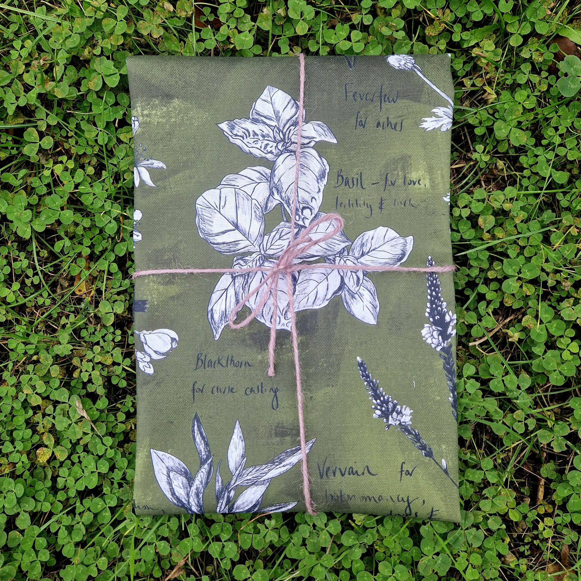 A folded green tea towel with white and black botanical plant illustrations, wrapped in lilac twine. The tea towel rests on a patch of green clover.