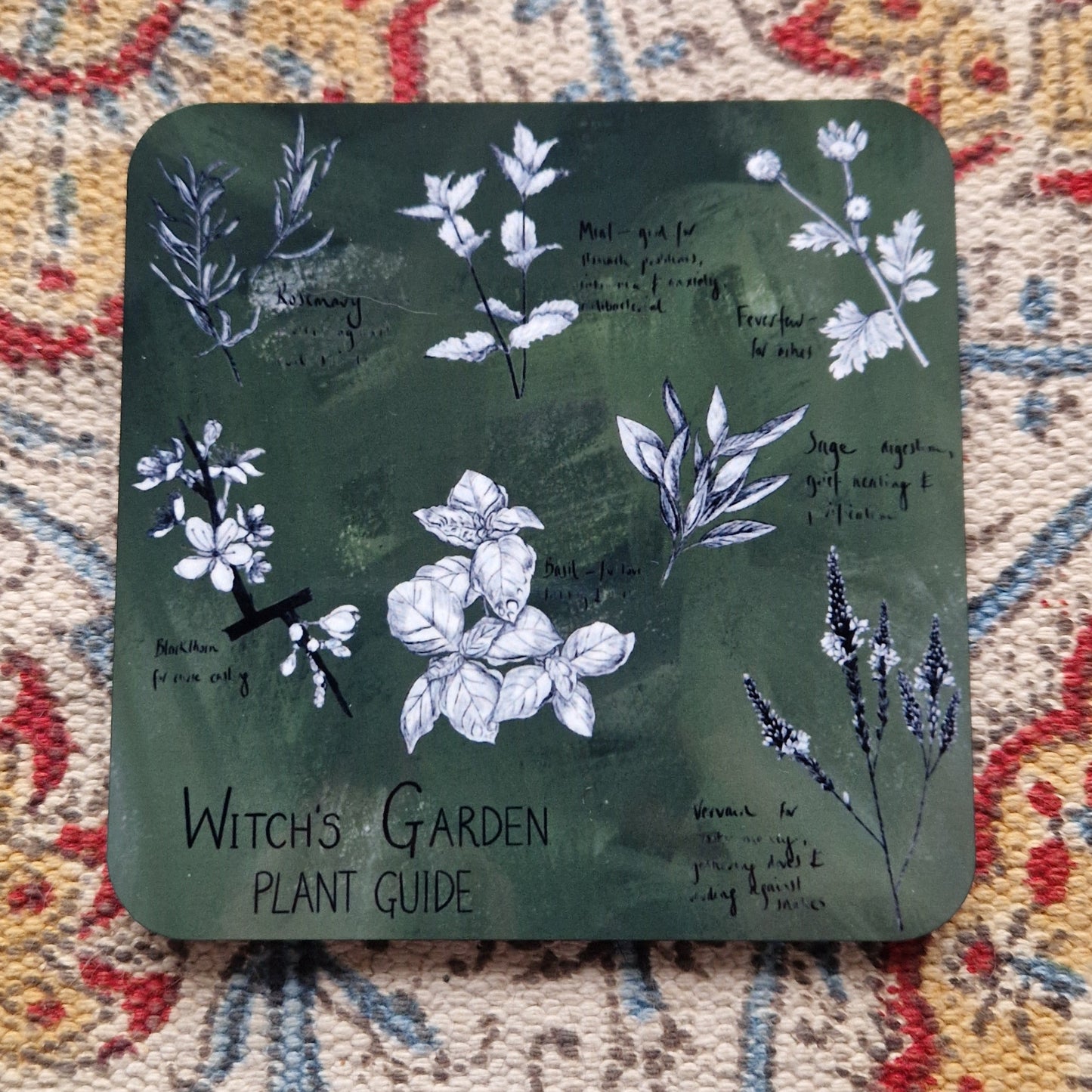 A green square coaster with a black and white illustrated plant design with the words 'Witch's Garden Plant Guide' at the bottom left in black handwriting.