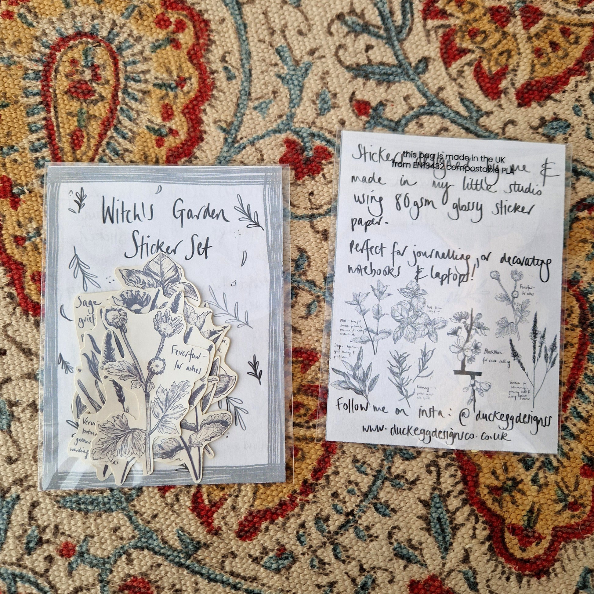 Two witch's garden sticker sets next to each other on a patterned fabric background. One front facing up showing the stickers inside the packaging and one back facing up showing the different designs inside the set.