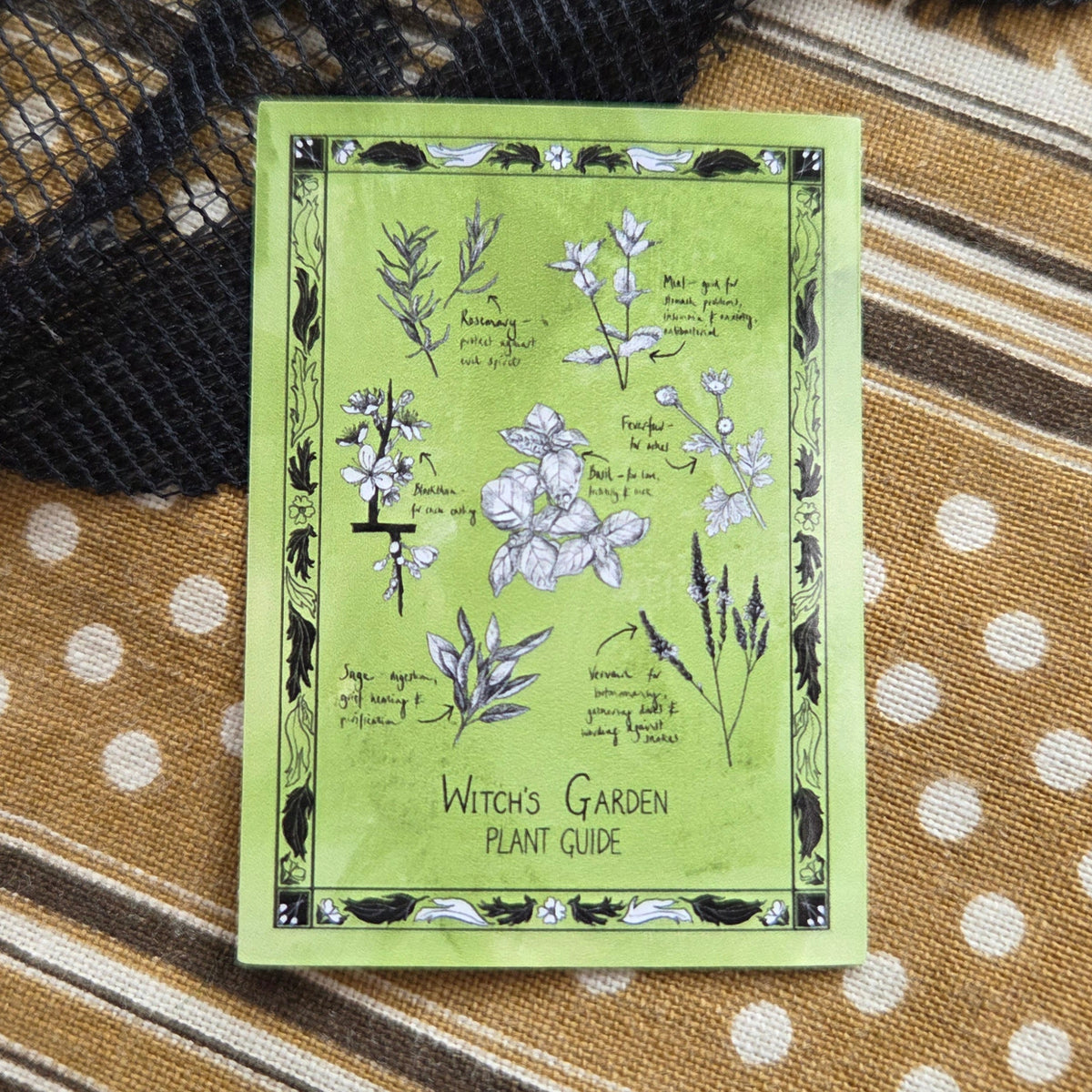 A vibrant green sticker with a black floral border and seven different plants you might find in a witchs garden. Behind the sticker is a brown patterned fabric with a black lace overlay.