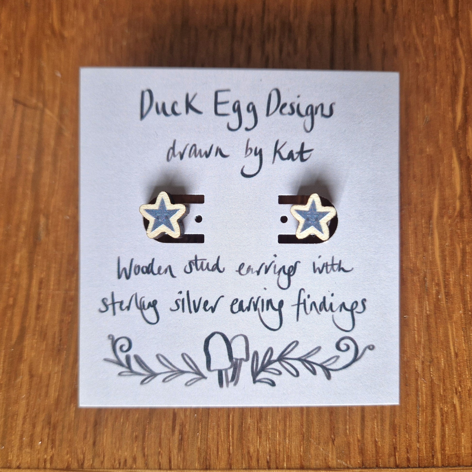 A pair of grey star earrings with small smiling faces sit on a light purple backing card with the words 'Duck Egg Designs drawn by Kat' across the top in black and 'Wooden stud earrings with sterling silver earring findings' across the bottom. Behind the backing card you can see a wooden background.
