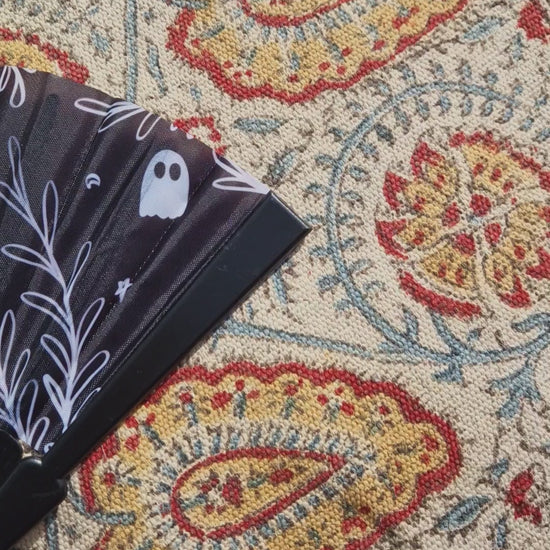 A video scanning across the fan from right to left. The fan is black with the words 'SPOOKY BITCH' in white across it alongside a leafy vine, ghost, crescent moon and stars design. The fan is resting on a patterned rug.