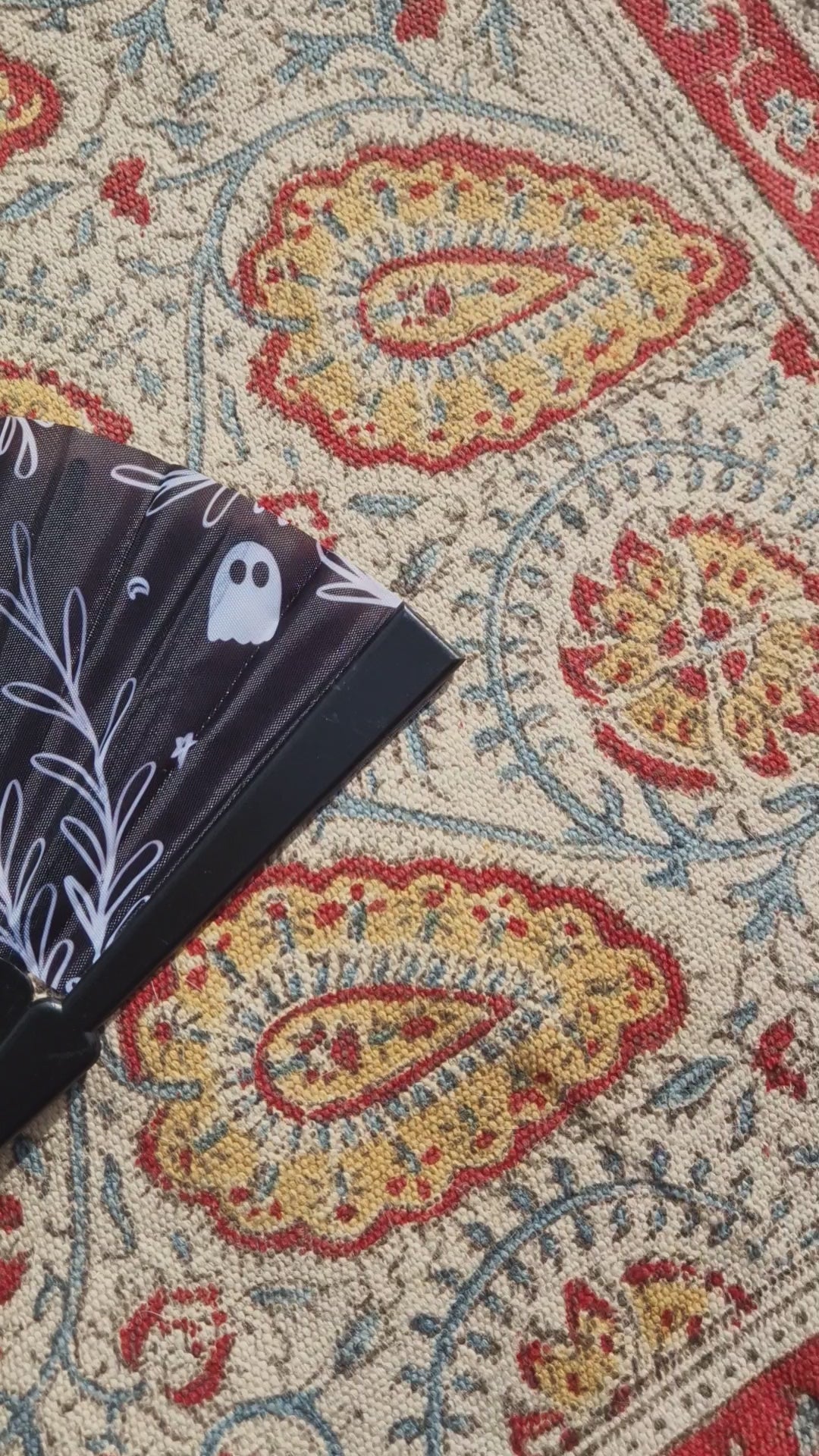 A video scanning across the fan from right to left. The fan is black with the words 'SPOOKY BITCH' in white across it alongside a leafy vine, ghost, crescent moon and stars design. The fan is resting on a patterned rug.