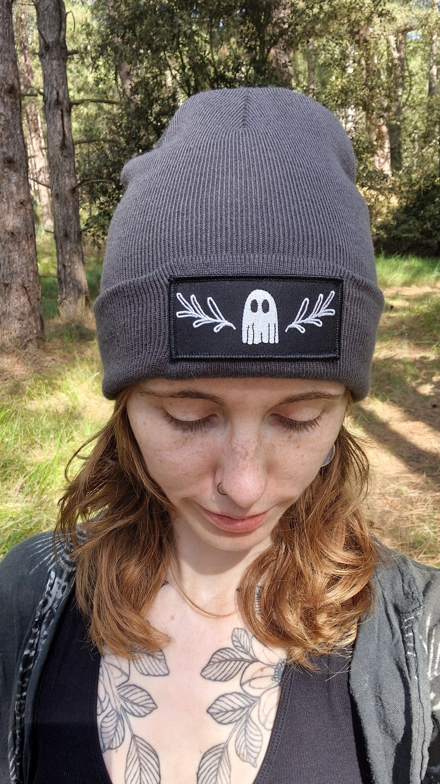 A person with light brown hair and chest tattoos stands in a forest, looking down while wearing Duck Egg Designs' Ghost Beanie—a gray recycled fabric hat with an iron-on white ghost and leafy branches patch.