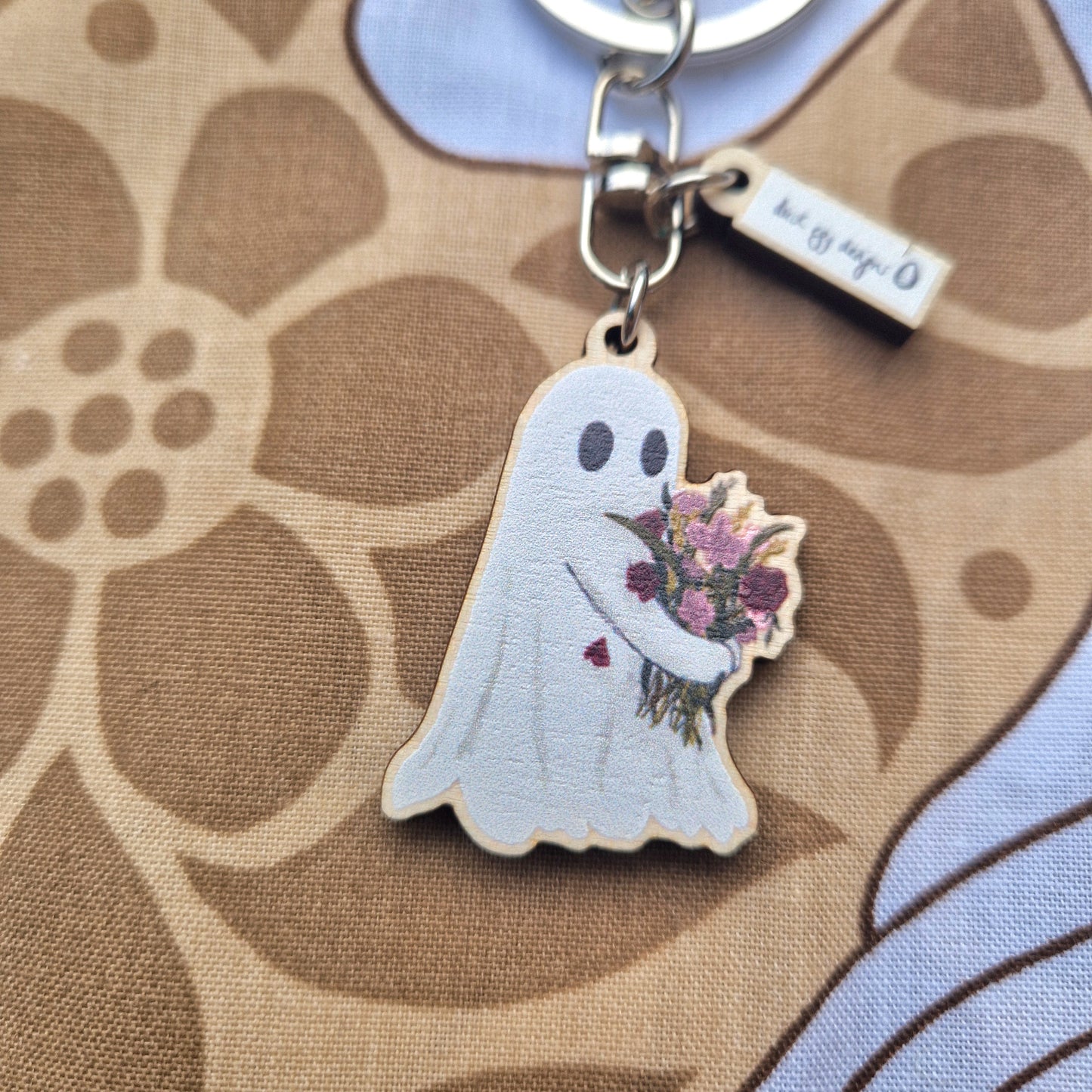 The Ghost Flower Keyring by Duck Egg Designs is a UK-made keychain shaped like a smiling ghost holding flowers, with a heart on its chest. Crafted from sustainable wood, it’s displayed on brown and beige floral fabric.