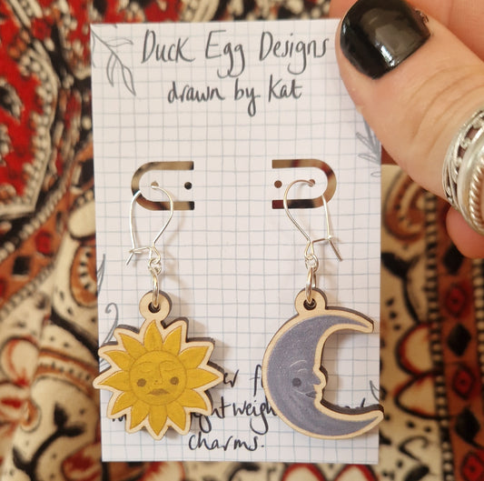 A hand displays a card reading Duck Egg Designs with Sun and Moon Face Earrings—one a smiling yellow sun, the other a cheerful crescent moon—charming wooden pieces by Kat.