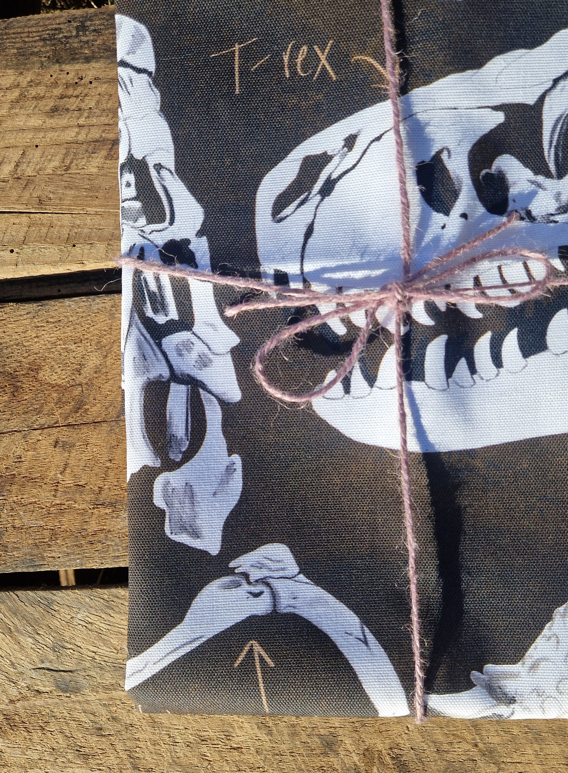 The Dinosaur Tea Towel by Duck Egg Designs is made from 100% cotton, features white T-rex skull illustrations, and comes folded with pink string on a wooden surface. An arrow highlights a bone on the fabric. Made in the UK.