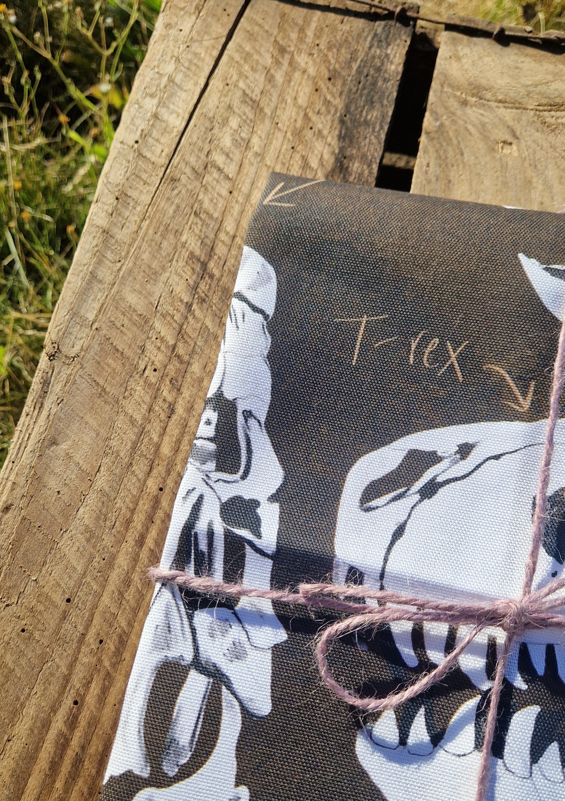 A close-up of a wooden surface with a package wrapped in Duck Egg Designs' Dinosaur Tea Towel, featuring dinosaur skull illustrations and handwritten T. rex labels, tied with pink string. Made in the UK from 100% cotton.