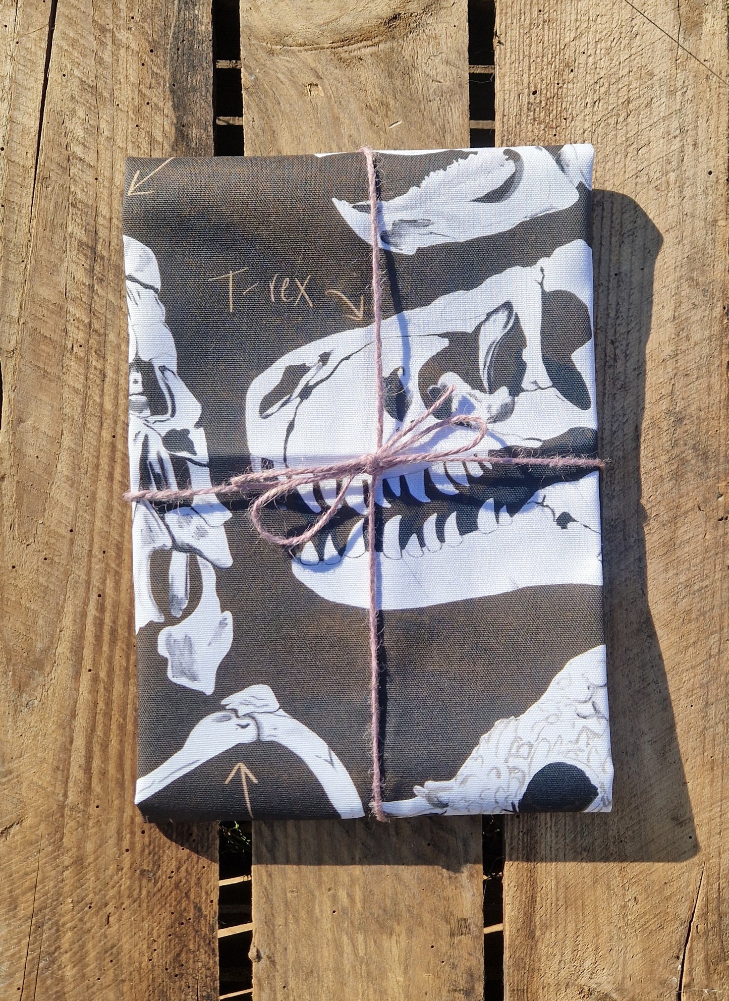 A gift wrapped in 100% cotton fabric featuring white T. rex skulls, tied with pink string, sits on a rustic wooden surface. Ideal for fans of Duck Egg Designs’ Dinosaur Tea Towel collection.