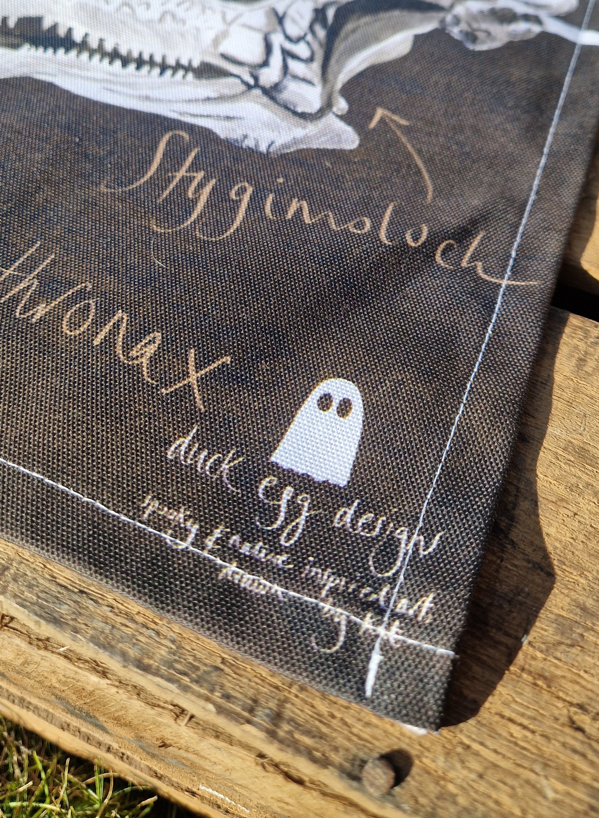 A close-up of the Dinosaur Tea Towel by Duck Egg Designs, made from 100% cotton with handwritten dinosaur names and a small ghost doodle, displayed on rustic wood. Made in the UK.