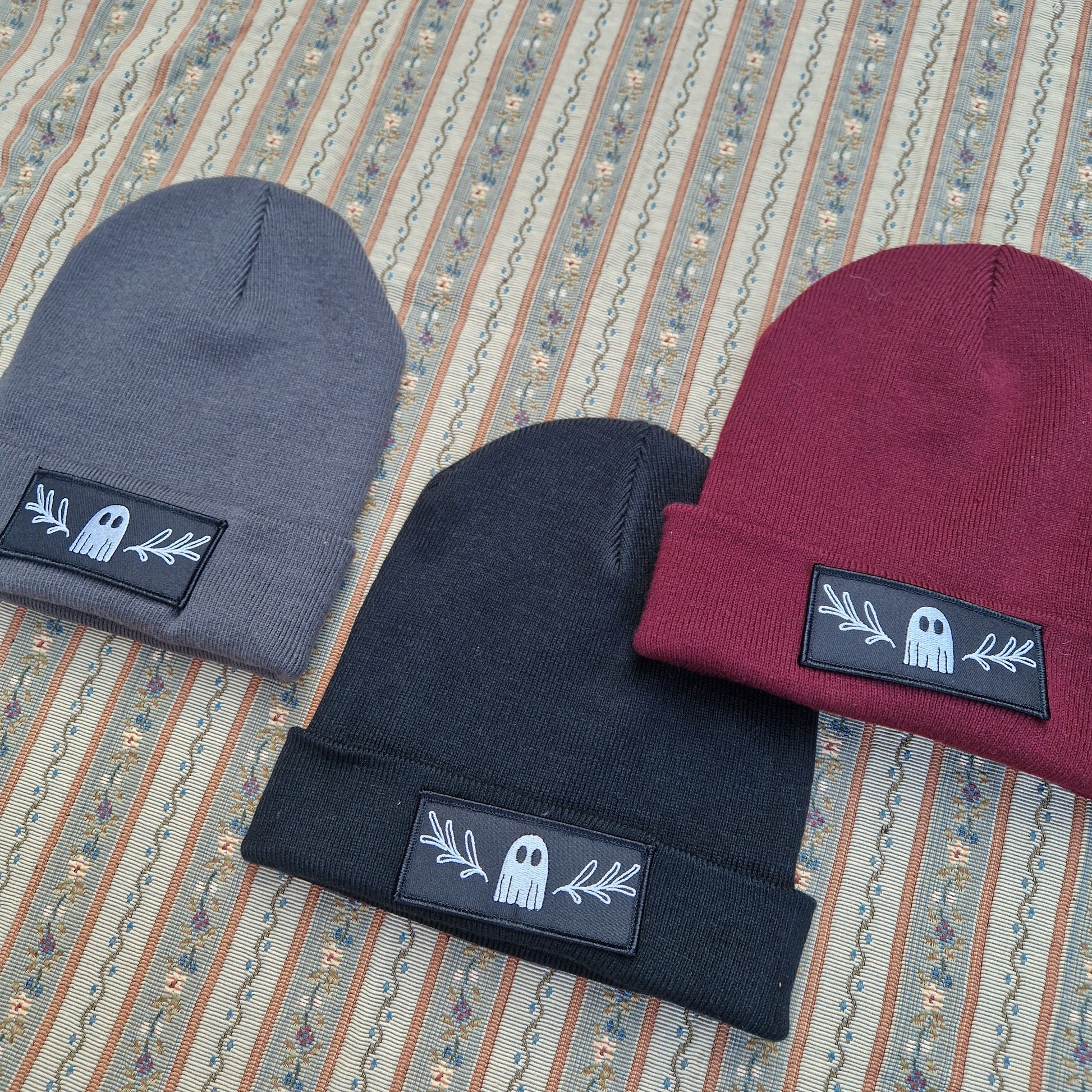 Three folded Duck Egg Designs Ghost Beanies—gray, black, and burgundy—each feature a patch with a white ghost and leaf design. They rest on a patterned fabric background with vertical stripes and small floral motifs.