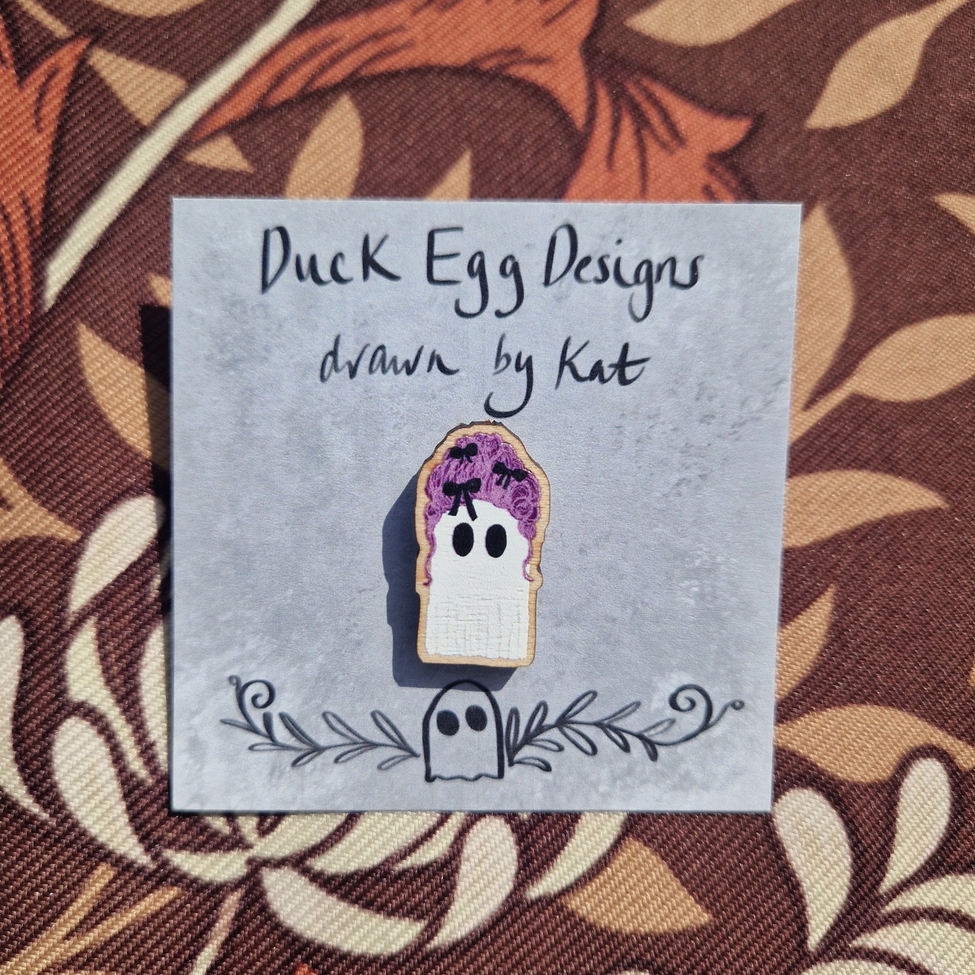 The Marie Antoinette Pink Wig Ghost Pin Badge by Duck Egg Designs is shown on a branded card, set against a brown and cream leafy fabric background.