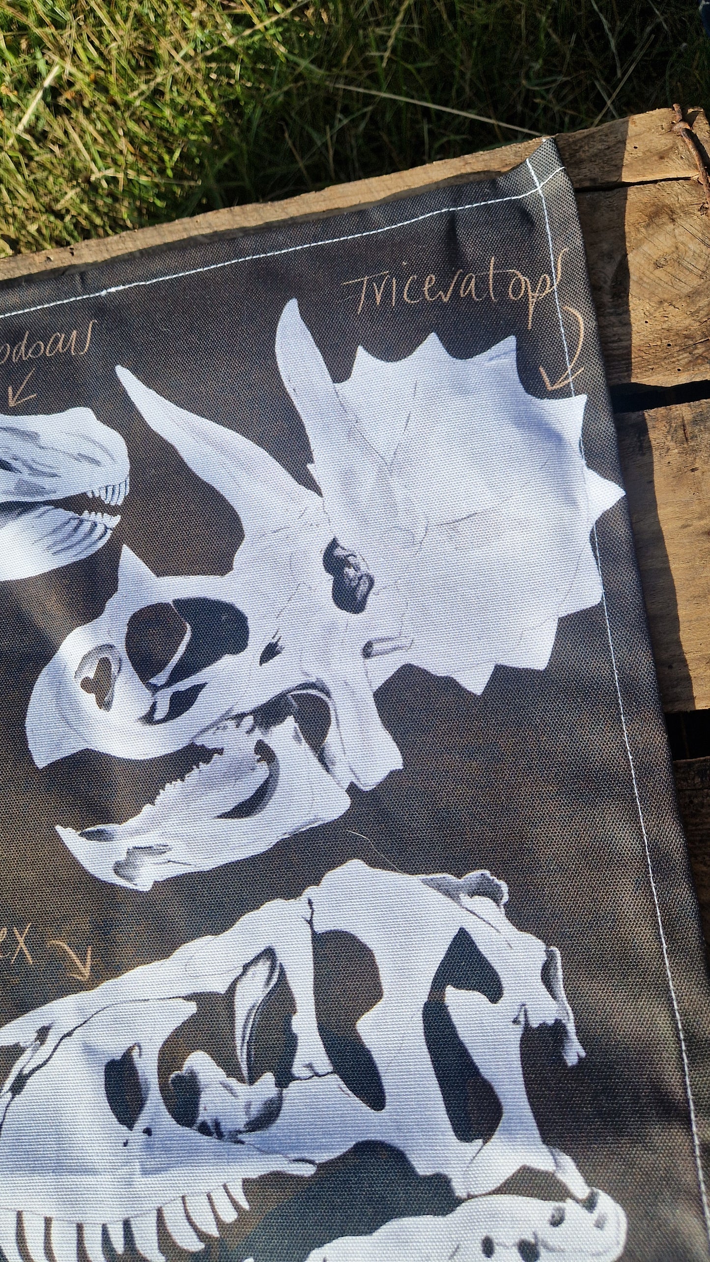 The Duck Egg Designs Dinosaur Tea Towel, made in the UK from 100% cotton, features white dinosaur skull illustrations—including a labeled triceratops—and is shown resting outdoors on wood with grass in the background.