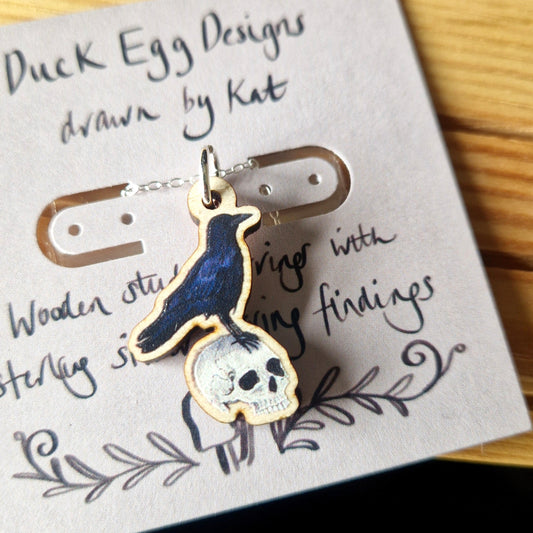 Crow on a human skull  jewellery pendant on a silver chain on a card with 'Duck Egg Designs' text.