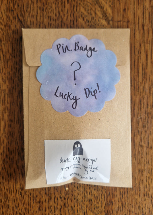A brown paper packet with a scalloped sticker reading "Mystery Pin Badge" and a Duck Egg Designs contact label hints at an enamel pin inside, resting on a wooden surface.