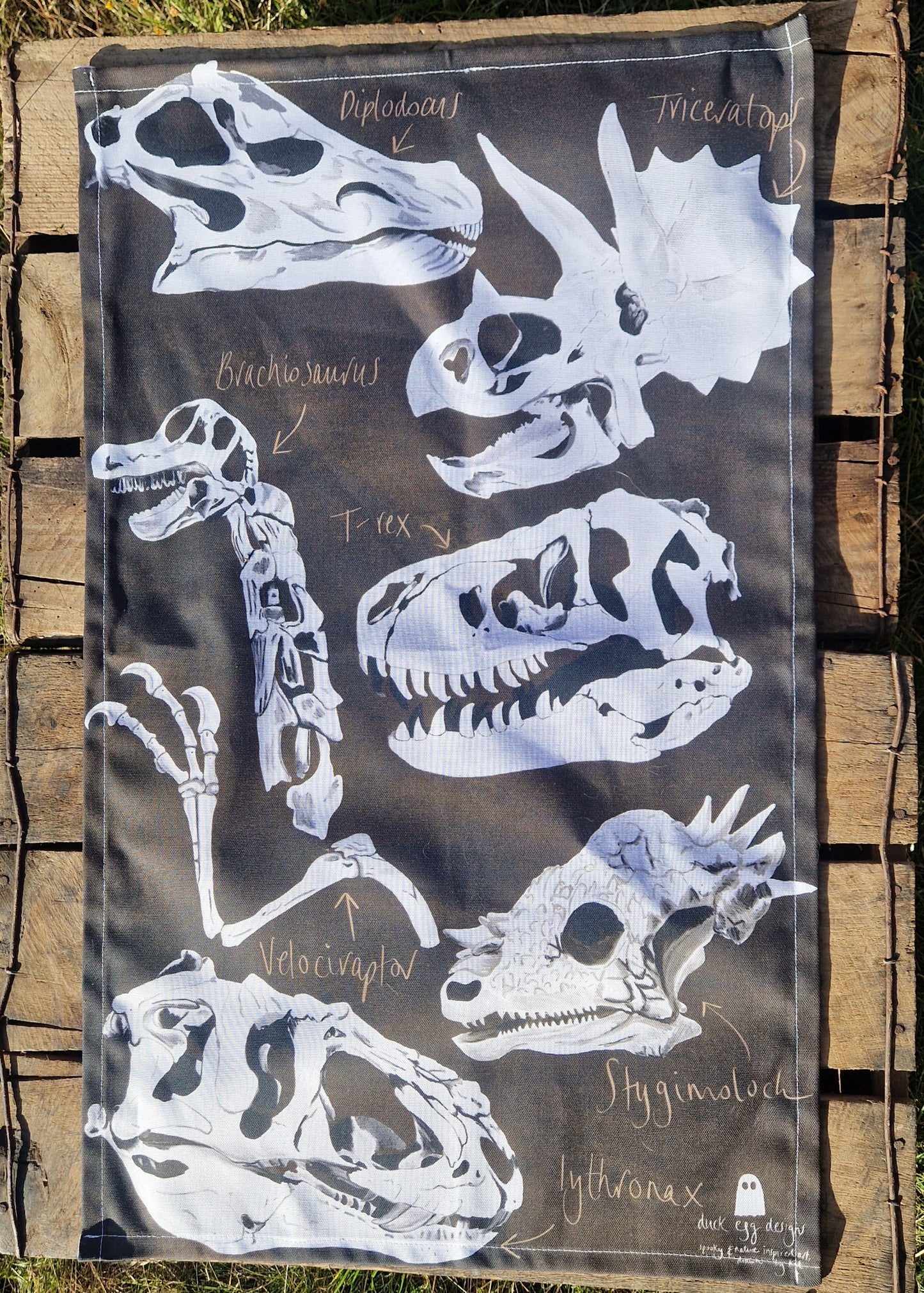The Dinosaur Tea Towel by Duck Egg Designs is 100% cotton, made in the UK, and features black-and-white labeled dinosaur skull illustrations like Diplodocus and Triceratops—shown here on wooden boards.
