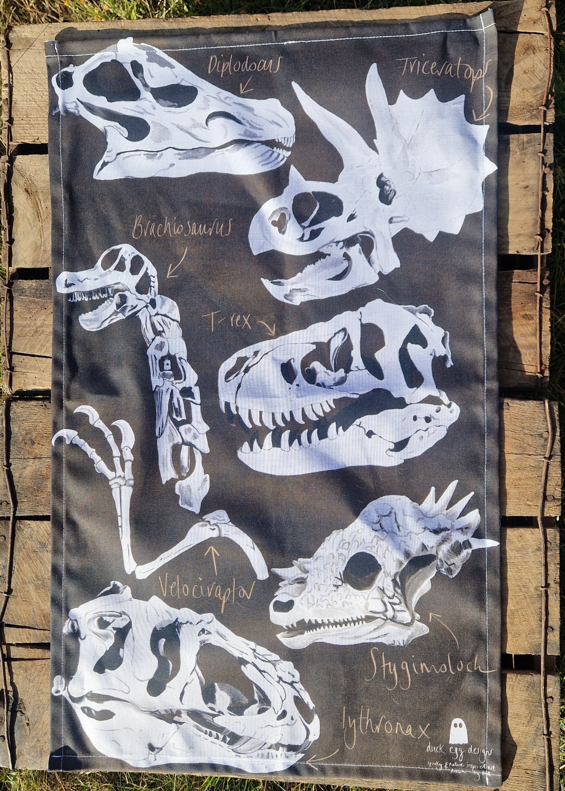 The Dinosaur Tea Towel by Duck Egg Designs is 100% cotton, made in the UK, and features black-and-white labeled dinosaur skull illustrations like Diplodocus and Triceratops—shown here on wooden boards.