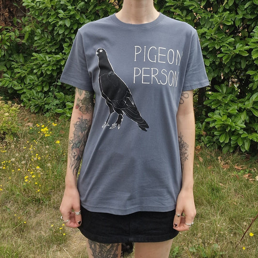 A tattooed person stands outdoors among green bushes and grass, wearing a gray Duck Egg Designs Pigeon Person Tshirt featuring a pigeon illustration and text, paired with a black skirt.