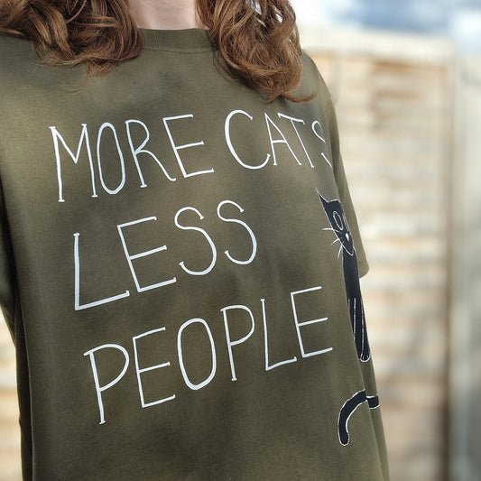 Outdoors in Norwich, a person with wavy brown hair wears the Duck Egg Designs More Cats Less People Tshirt featuring a black cat drawing. A wooden fence and cloudy sky add to the cozy vibe.