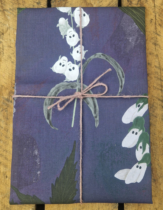 A rectangular gift wrapped in purple fabric with a white lily of the valley pattern and twine bow rests on rustic wood—an ideal match for Duck Egg Designs Spooky Flowers Tea Towel.