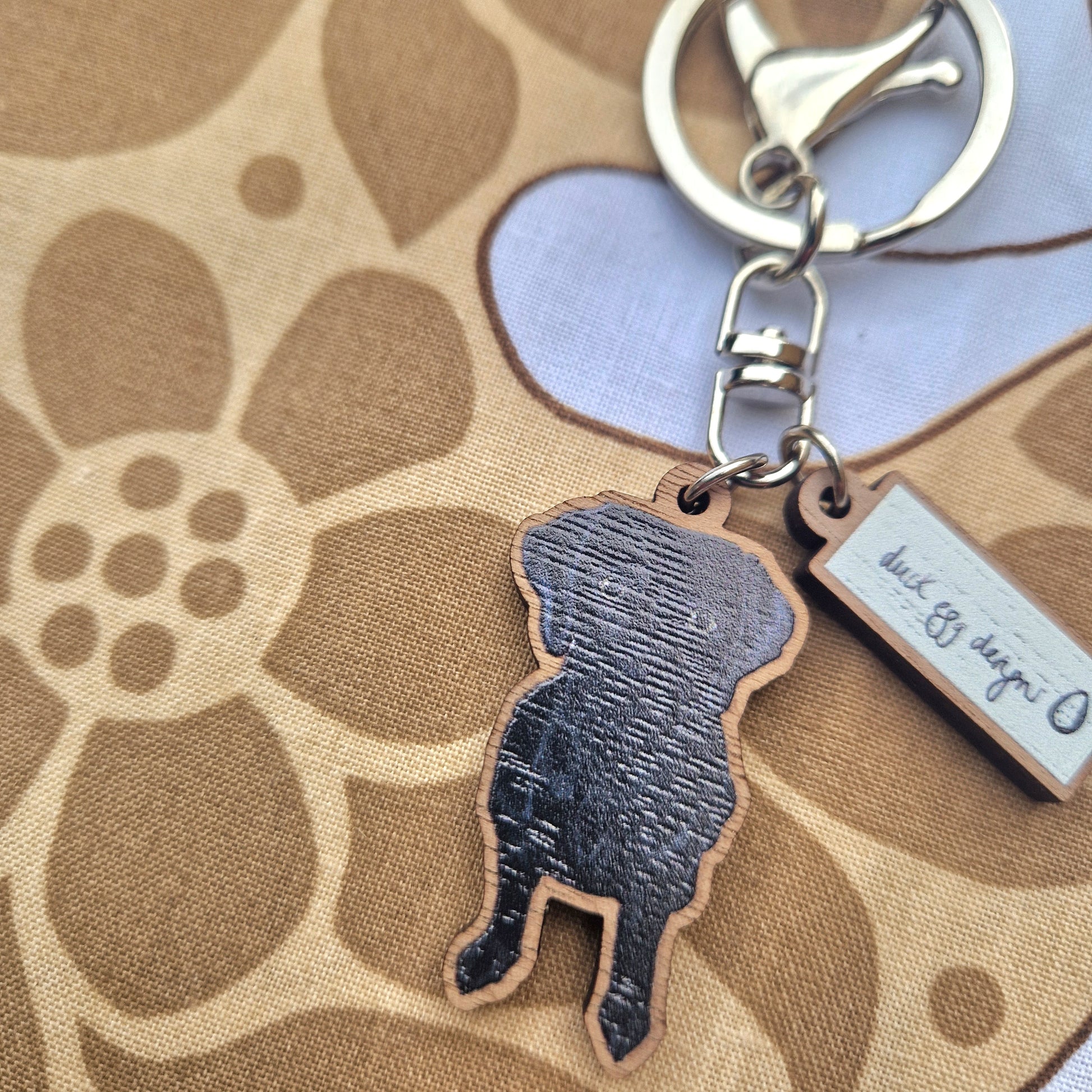 The Rondo Black Labrador Keyring by Duck Egg Designs, featuring a black labrador charm and small rectangular tag on a metal keyring, rests on brown and white floral-patterned fabric.