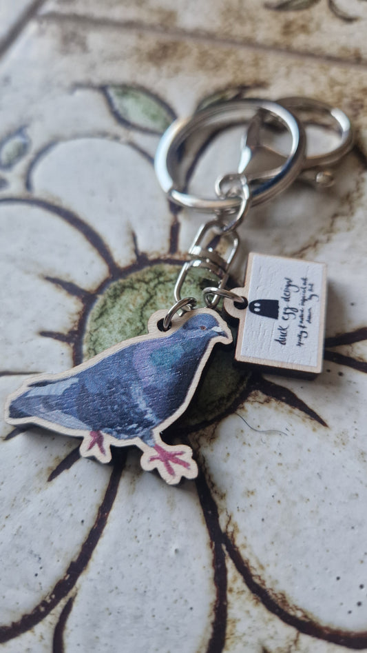 The Paloma Pigeon Keyring by Duck Egg Designs features a wooden pigeon charm and a small rectangular tag with handwritten text and a pen drawing, displayed on a floral tile.