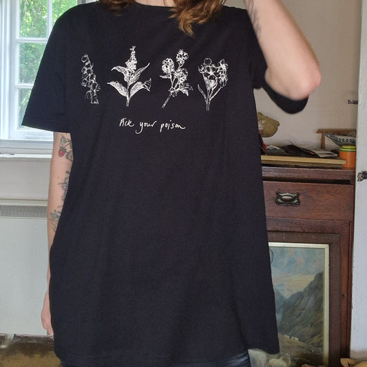A woman with light brown hair wears the Duck Egg Designs Black Poison Plants Tshirt, a loose black unisex shirt with white botanical prints and "Pick your poison" text. She stands indoors with one hand near her ear.