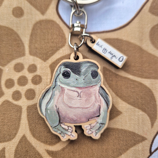 The Duck Egg Designs Frog Keyring is a sustainable wood keychain featuring a cute, big-eyed frog with a light purple belly, attached to a metal ring and small tag, set against a brown floral background.