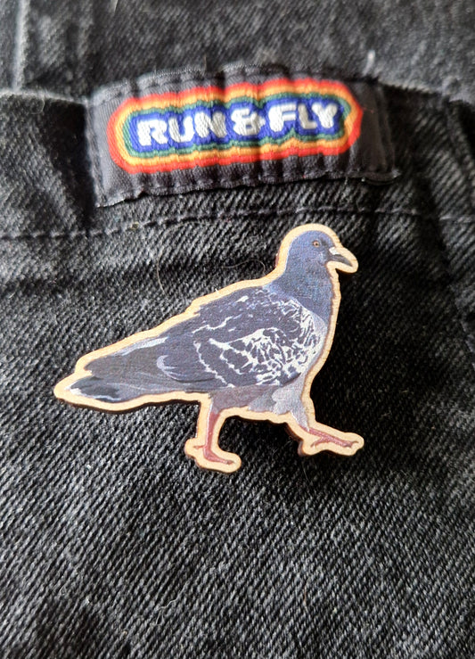 The Cyril Pigeon Pin Badge by Duck Egg Designs is made in the UK and attached to black denim fabric beneath a colorful RUN & FLY label.