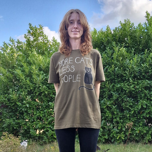 In Norwich, someone smiles by green bushes wearing the Duck Egg Designs "More Cats Less People" Tshirt with a cat print. A black and white cat lounges in the grass nearby under a partly cloudy sky.