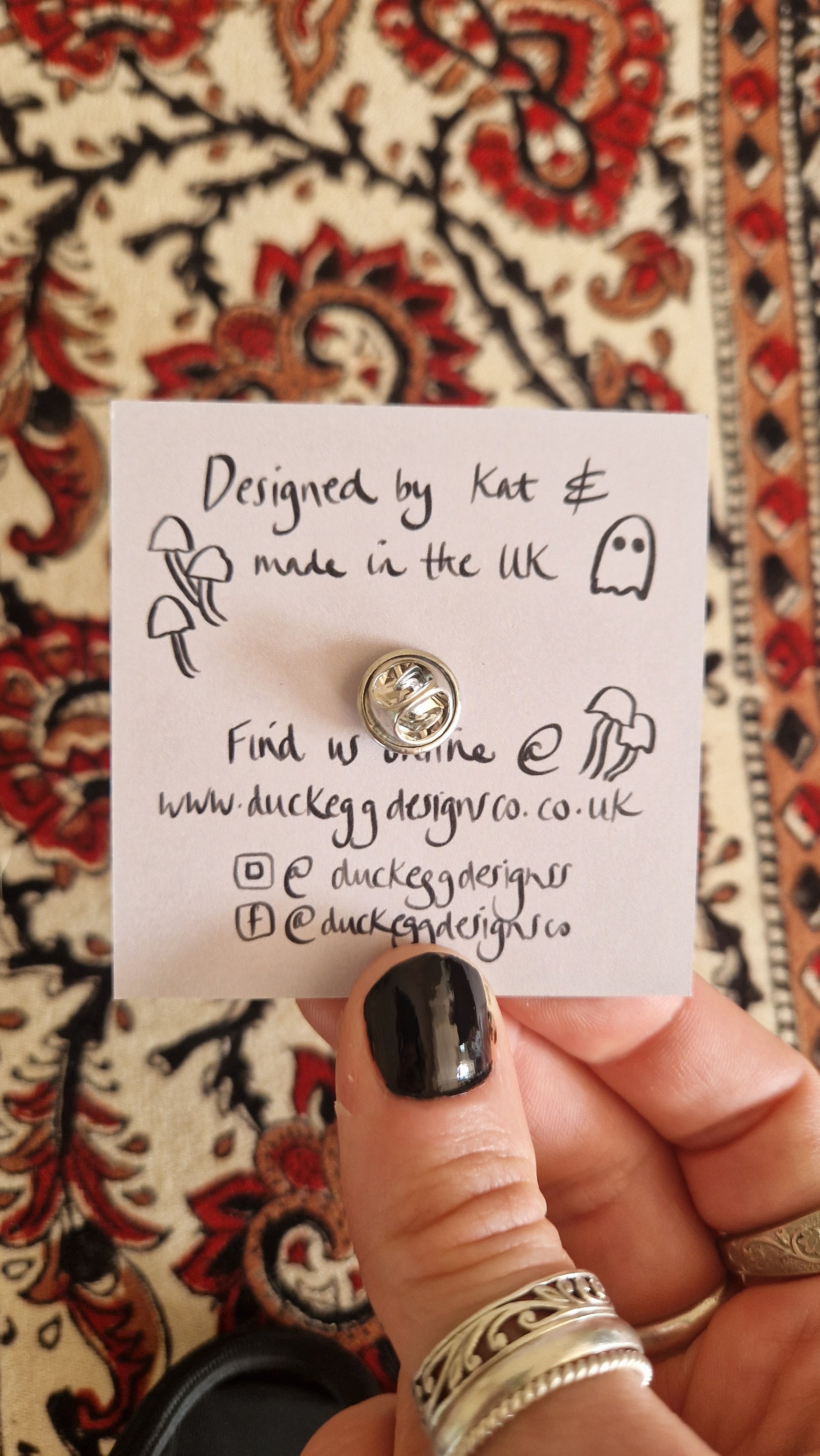 A hand with black nail polish holds a Marie Antoinette Pink Wig Ghost Pin Badge by Duck Egg Designs, attached to a card with mushroom and ghost doodles plus social info for duckeggdesignco.co.uk, over patterned fabric.