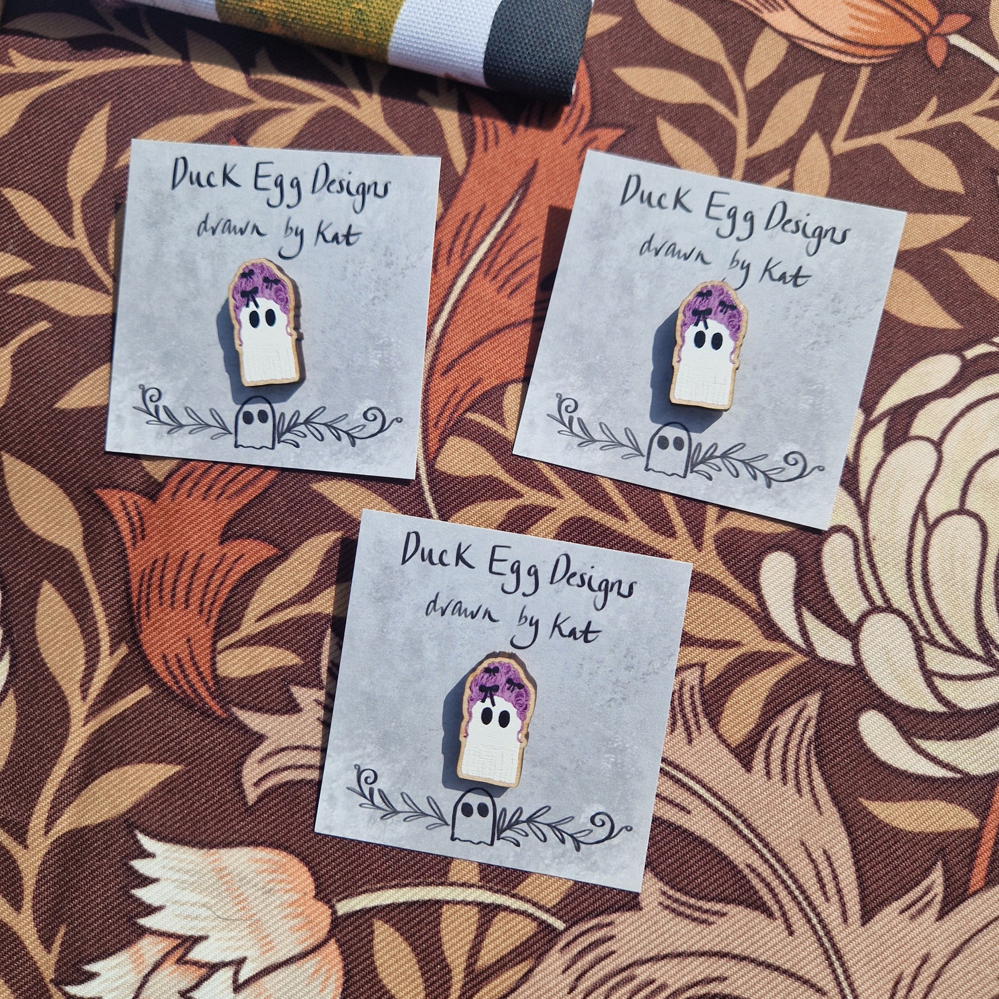 Three Marie Antoinette Pink Wig Ghost Pin Badges by Duck Egg Designs are displayed on branded cards, their charming design with purple flowers adding a whimsical touch to any collection.