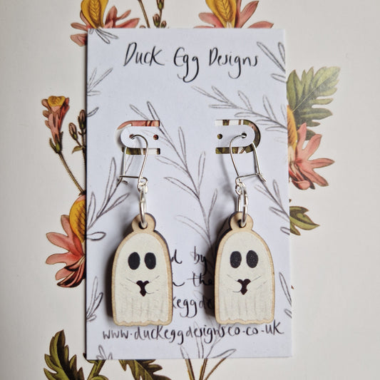 Ghost With Heart Earrings by Duck Egg Designs feature wooden ghost charms with heart cutouts, black eyes and mouths, displayed on a branded card with floral art. A playful accessory to enhance any outfit.