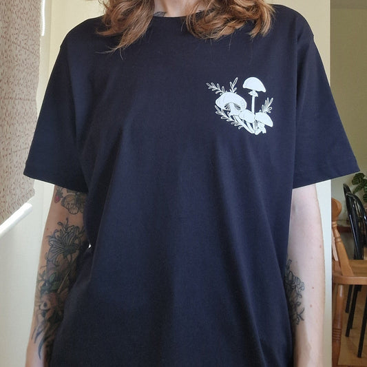 A person wears the Duck Egg Designs Black Fungi Tshirt With Back And Front Print, showing a white mushroom and leaf graphic on the left chest. Their floral tattooed forearms are visible in front of a dining area and window.