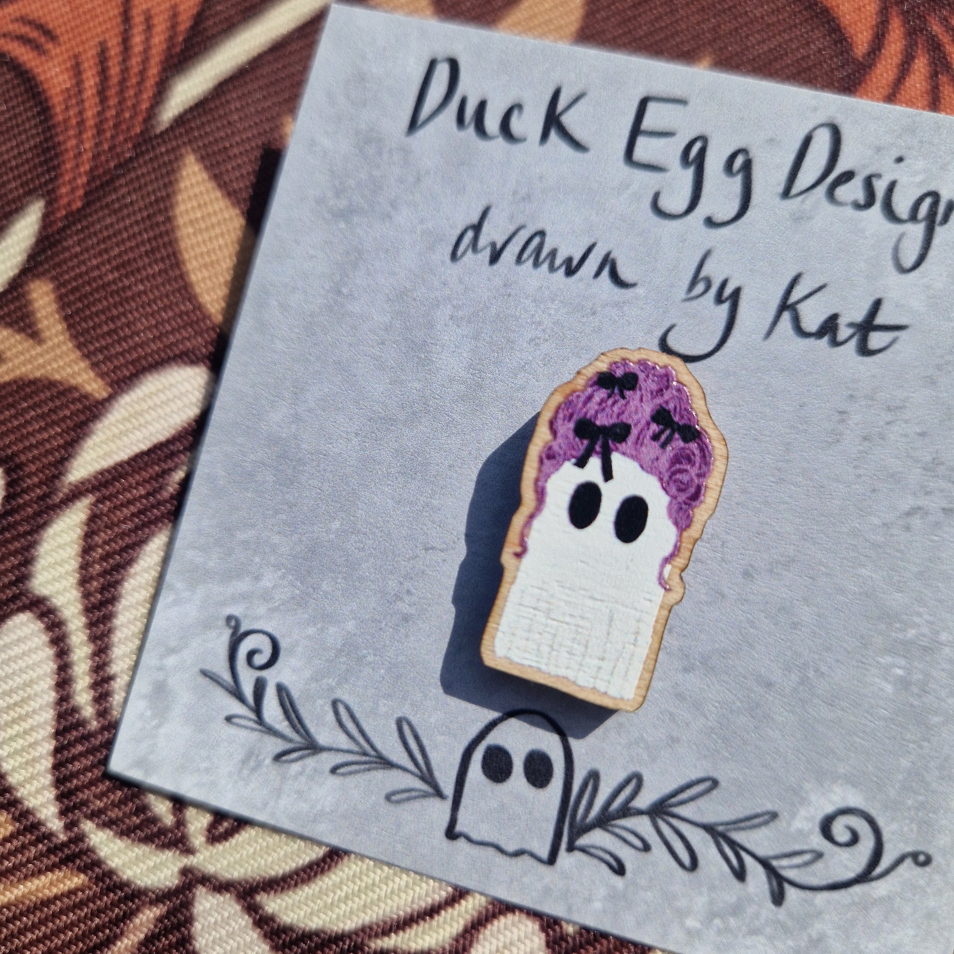 The Marie Antoinette Pink Wig Ghost Pin Badge by Duck Egg Designs is a wooden ghost pin with a pink wig, attached to a branded card illustrated by Kat—perfectly spooky and unique!.